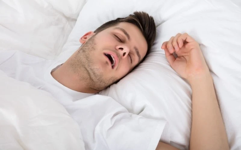 Illustratives Bild zum Thema Schnarchen: Ein junger Mann mit leicht geöffnetem Mund liegt schlafend im Bett.